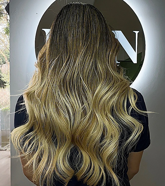 Balayage rubio largo con ondas, de espaldas frente al letrero iluminado DAN